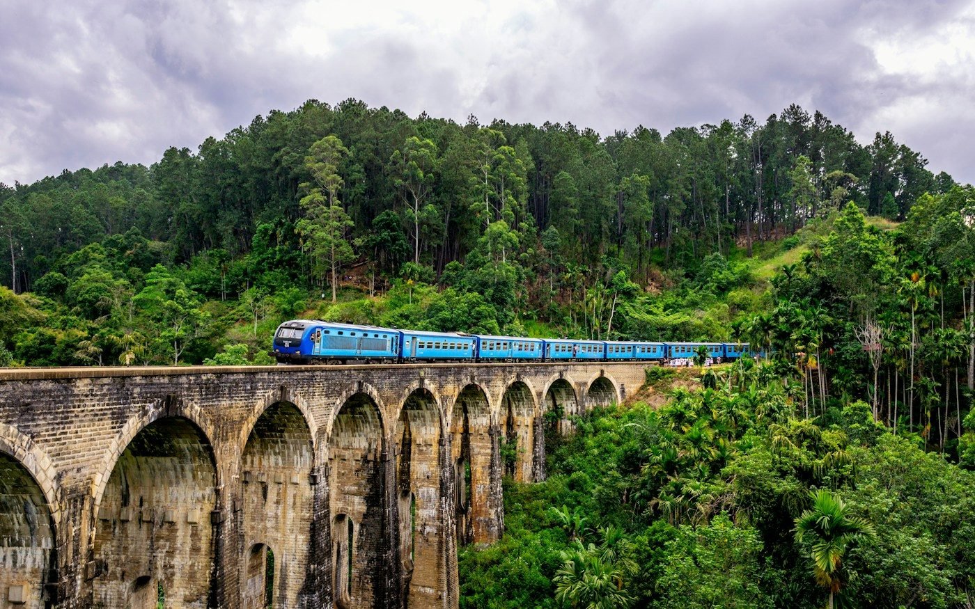 A blue train crossing the Nine Arch Bridge in Ella, surrounded by forest